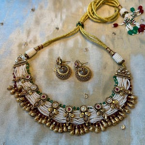 May include: A gold-toned necklace and matching jhumka earrings set featuring multiple strands of small white beads, accented with green and red stones, gold metal floral motifs, and small dangling gold charms on a gold cord.