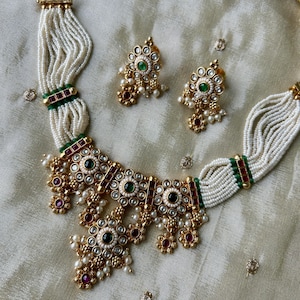 May include: A gold-toned necklace and matching earring set featuring multiple strands of small white seed beads, accented with green and red stones, clear crystals, and dangling pearl-like beads. The necklace has a central ornate pendant.
