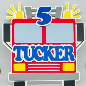 May include: Red fire truck cake topper with a blue number 5 and the name Tucker in blue letters. The fire truck has yellow lights and a gray ladder.