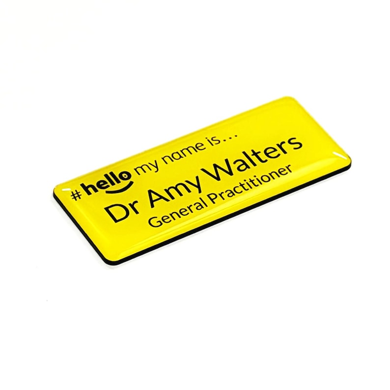 May include: A yellow name tag with black text. The tag reads "#hello my name is... Dr Amy Walters General Practitioner". The tag has a black border and is slightly angled.