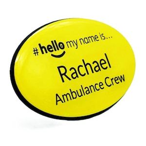 May include: A yellow oval name badge with a black border. The badge reads "#hello my name is..." followed by the name "Rachael" and "Ambulance Crew" in black text. The badge has a glossy finish.