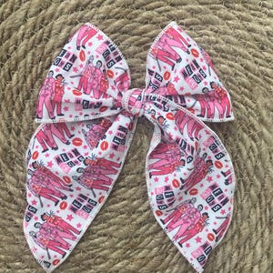 May include: A large, white hair bow with a repeating pattern of pink figures, red lips, and stars. The text "MEAN BOYS" is visible on the bow. The bow is tied in the center and has long, flowing tails.