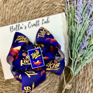 May include: A large, handmade hair bow featuring a vibrant blue ribbon with a repeating "Takis" and flame design. A small "Takis" logo charm is centered on the bow. The bow is set against a white card with the text "Bella's Craft Lab".