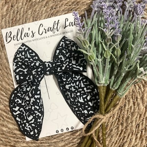 May include: A black and white patterned hair bow with a matching bow tie is displayed on a card that reads "Bella's Craft Lab". A bundle of lavender stems is tied with twine, adding a touch of color to the scene.