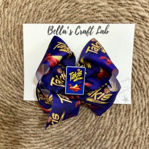 May include: A large, handmade hair bow featuring a repeating pattern of the Takis logo and red and yellow graphics on a navy blue background. A small Takis logo charm is centered on the bow. The bow is attached to a white card with the text "Bella's Craft Lab".