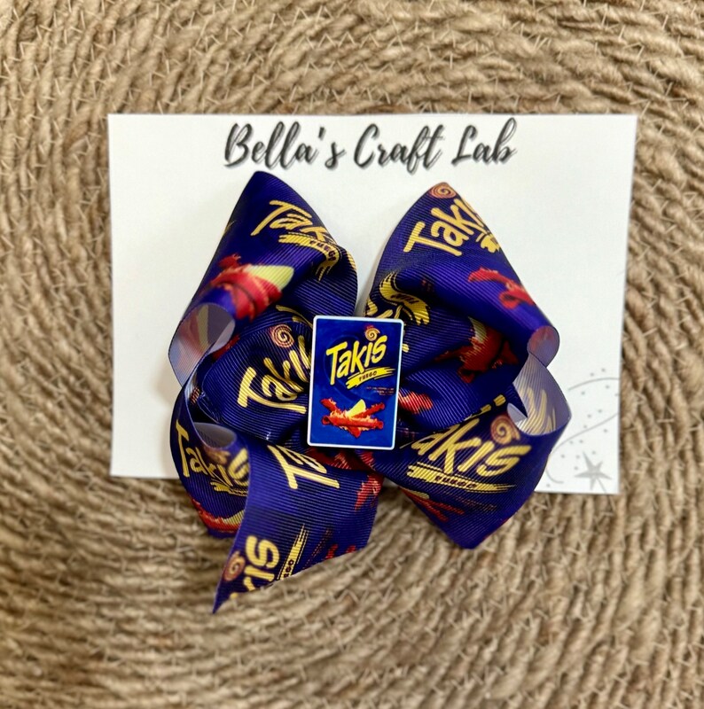 May include: A large, handmade hair bow featuring a repeating pattern of the Takis logo and red and yellow graphics on a navy blue background. A small Takis logo charm is centered on the bow. The bow is attached to a white card with the text "Bella's Craft Lab".