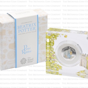 Puede incluir: Una moneda de plata con Peter Rabbit de la serie de libros infantiles de Beatrix Potter. La moneda se muestra en una caja de acrílico transparente con una ilustración colorida de Peter Rabbit en un jardín. La caja está etiquetada con "Celebrating Beatrix Potter and Her Little Tales" y "The Royal Mint".