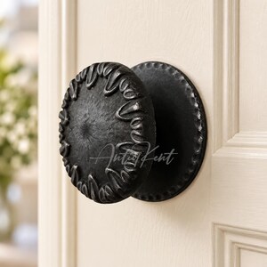 May include: A round, black metal door knob with a textured, scalloped edge and a matching circular backplate. The knob features a dark, matte finish with a raised, petal-like pattern around the perimeter. The text AntiqKent is visible.