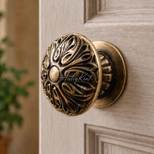 May include: A round brass door knob with an intricate, embossed floral pattern and a smooth central circular disc. The metal has a dark, aged patina finish with highlights on the raised edges. The text AntiqKent is visible on the knob surface.