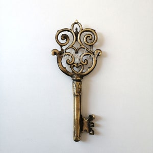 May include: A decorative ornate key made of gold metal with a detailed design. The key has a large, intricate head and a long, slender shaft.