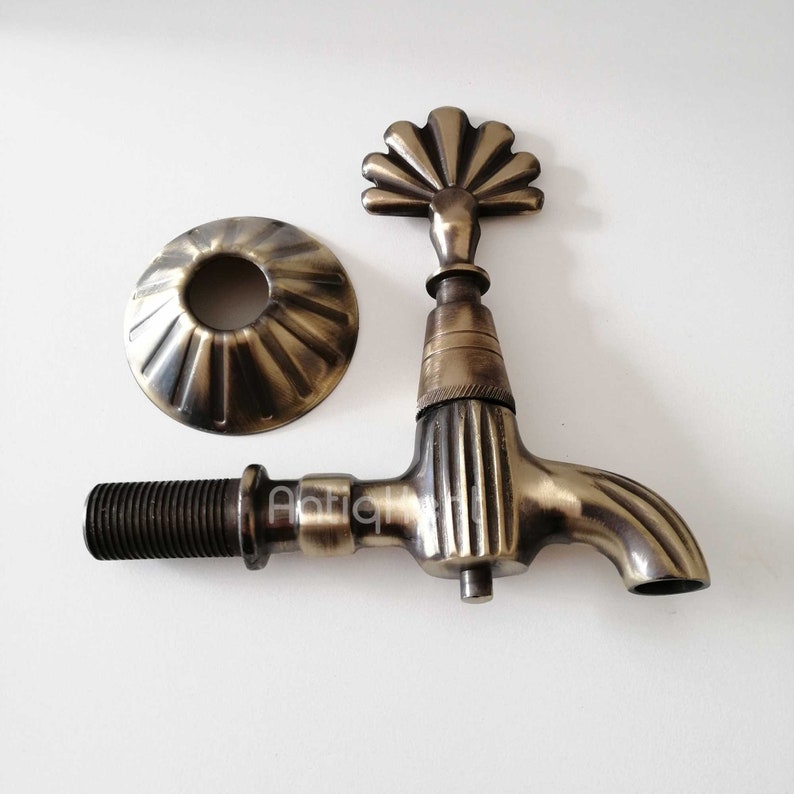 May include: Antique brass faucet with a decorative fan-shaped handle and a round base. The faucet has a ribbed design and a spout that curves to the side.