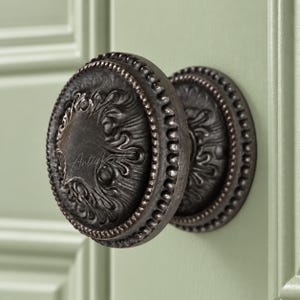 May include: A round, dark bronze-toned metal cabinet knob featuring an intricate, raised relief pattern on its face and a beaded border. The knob is mounted on a light green paneled surface and displays the text "AntiqKent" in a subtle script.