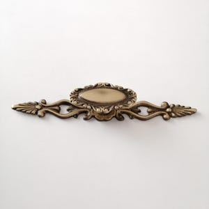 May include: A gold-toned metal drawer pull with an oval-shaped centre and ornate scrollwork on either side.