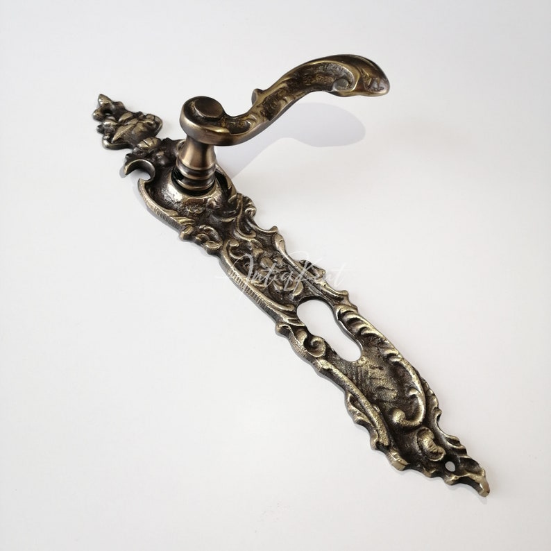 May include: Ornate brass door handle with a keyhole and a decorative backplate. The handle is curved and has a detailed design.