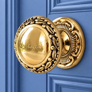 Könnte beinhalten: Ein polierter goldfarbener Messing-Türknauf mit einer glatten, reflektierenden Mitte und einem dekorativen Rand mit geprägten Blatt- und Beerenmotiven. Das Wort Antig Kent ist in der Mitte des Knaufs eingraviert, der an einer blauen Tür montiert ist.