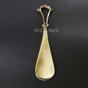 May include: A gold-toned shoehorn with an ornate, floral design on the handle. The handle is shaped like a loop and the shoehorn has a long, narrow blade.