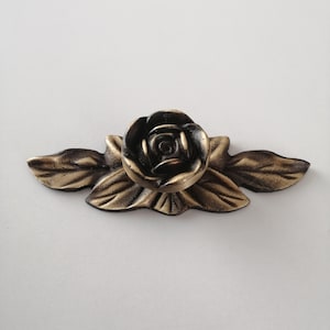 May include: A gold-toned metal cabinet knob with a rose design. The rose is black and has a detailed design. The leaves are gold-toned and have a textured surface.