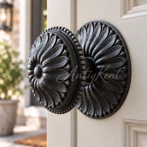 May include: A dark, metallic, circular door knob featuring a raised, radiating petal or floral pattern. The knob has a textured, ridged edge and is mounted on a matching circular backplate with the same petal design, attached to a light-colored door.