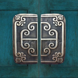 Puede incluir: Un par de tiradores de puerta de plata adornados con un diseño de desplazamiento decorativo. Los tiradores están montados en una puerta de madera azul turquesa.