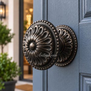 May include: A round, dark bronze-toned metal door knob featuring a raised, swirling petal-like pattern and a beaded outer edge. The knob is mounted on a matching circular backplate and is attached to a dark blue, paneled wooden door. The text AntigKent is visible.