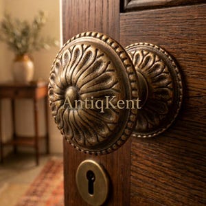 Puede incluir: Un pomo de puerta redondo de latón antiguo con un patrón detallado de pétalos radiantes y una placa de cerradura circular a juego, montado en una puerta de madera oscura y texturizada. El texto AntiqKent está superpuesto sobre el pomo.