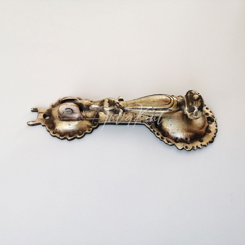 May include: A vintage brass door knocker with an ornate design. The knocker has a rounded handle and a decorative plate with a scalloped edge.