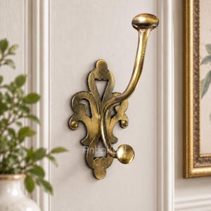 May include: A wall-mounted coat hook with an ornate, openwork metal backplate in a distressed brass finish. The hook features a long, curved upper arm and a shorter, rounded lower arm, secured to the wall with two visible brass-colored screws.