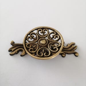 May include: Ornate brass-toned cabinet pull with a circular, filigree design. The center features a small, raised circle, and the pull extends with decorative, leaf-like elements on each side. A vintage-style hardware piece.