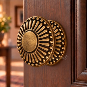 Peut inclure: Un bouton de porte rond en laiton antique doté d'un disque central plat entouré d'un motif rayonnant de crêtes en relief et de petites perles sphériques, fixé sur un panneau de porte en bois brun foncé avec un grain visible.