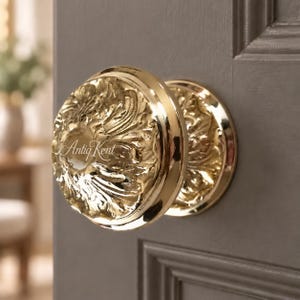 May include: A polished gold-tone round door knob featuring an intricate embossed floral and leaf pattern across its surface. The knob is mounted on a dark grey panelled door and displays the text "AntiqKent" in a white script font on its face.