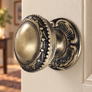 Puede incluir: Un pomo redondo para mueble con acabado en latón antiguo, que presenta un domo central liso rodeado por un borde decorativo con intrincados detalles en relieve. El pomo está montado sobre una placa circular con un borde festoneado a juego.