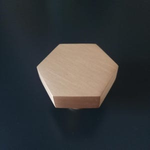 Puede incluir: Un objeto hexagonal de madera de color marrón claro. El objeto tiene una parte superior plana y lados angulados, con una textura de veta de madera visible. El objeto está sobre un fondo oscuro.
