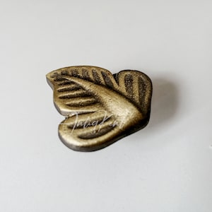 May include: A bronze-coloured leaf-shaped cabinet pull with detailed veining. The hardware has a distressed finish and is likely made of metal. The text "Antiq Kent" is visible on the pull.