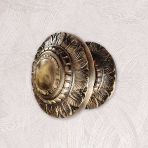 Może przedstawiać: Dwa antyczne mosiężne gałki drzwiowe z misternymi wzorami kwiatowymi. Gałki są okrągłe i mają wypukły środek.