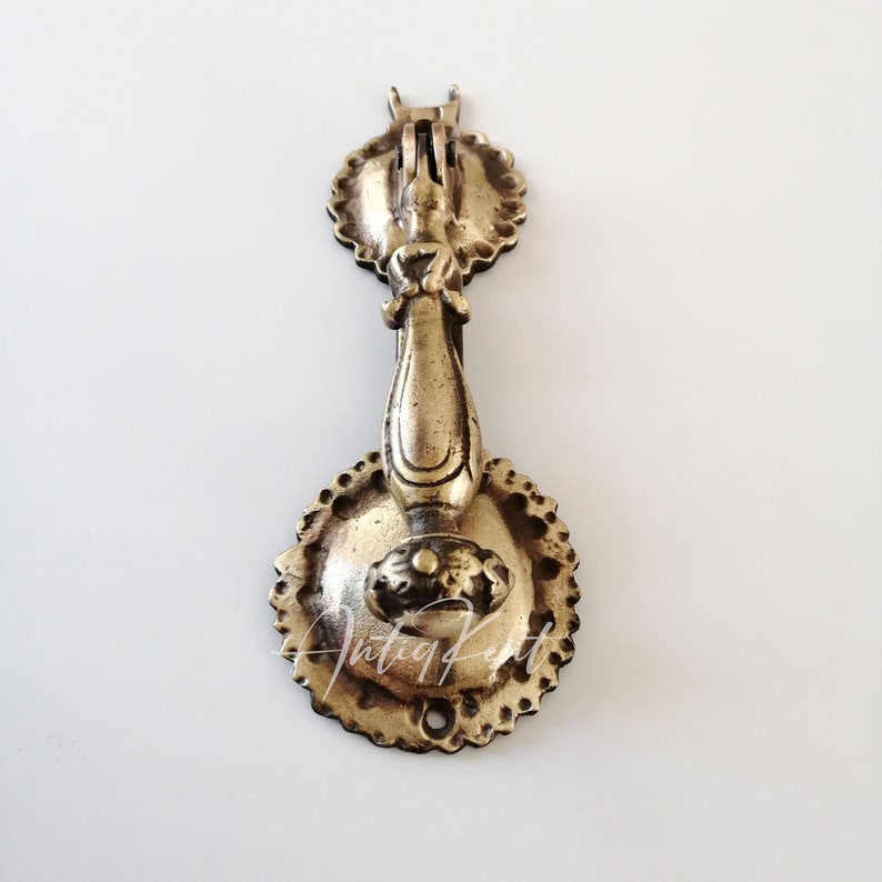 May include: Antique brass door knocker with a decorative floral design. The knocker has a round base and a long, ornate handle.