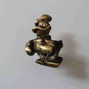 May include: A small, brass-colored figurine of Donald Duck, a cartoon character. The figurine is in a seated position, wearing a sailor suit and hat. The figure has a detailed design and is on a white background.