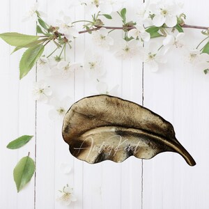 May include: A gold leaf-shaped dish on a white wooden background. The dish is made of metal and has a textured surface.