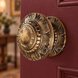 May include: A round brass door knob with a detailed leaf-like pattern radiating from a smooth central disc. The knob features an antique finish with dark highlights in the crevices and is mounted on a deep burgundy door with a panel design.