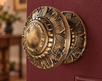 May include: A round brass door knob with a detailed leaf-like pattern radiating from a smooth central disc. The knob features an antique finish with dark highlights in the crevices and is mounted on a deep burgundy door with a panel design.