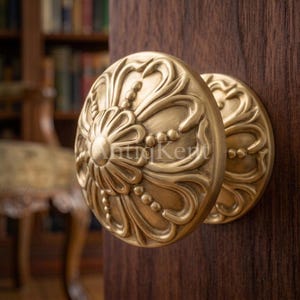 Peut inclure: Un bouton de meuble rond en laiton orné d'un motif floral en relief complexe avec des détails perlés. La quincaillerie présente une finition métallique brossée et est fixée sur une surface en bois brun foncé, révélant un design symétrique.