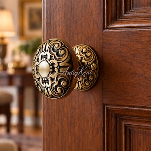 May include: A round brass door knob with an intricate embossed scrollwork pattern and a smooth central circular plate, mounted on a dark wood door. The text AntiqKent is visible in white script across the center of the metal door knob surface.