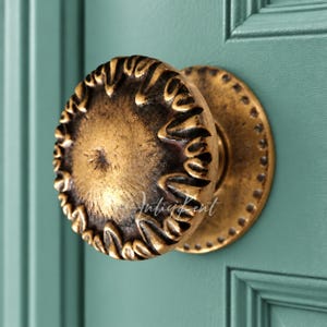 May include: A round, antique brass-finished door knob with a textured, scalloped edge and a central dimple. The knob features a dark, weathered patina and is mounted on a circular backplate with a decorative beaded border detail.