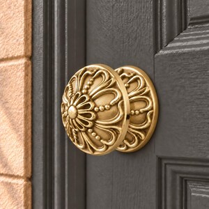 May include: A round, metallic gold door knob featuring an intricate, raised floral pattern with symmetrical petals and beaded detailing. The knob is mounted on a dark gray, paneled door, showcasing a polished finish with subtle highlights.