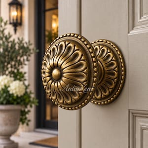 May include: A round brass door knob with a raised floral petal pattern and a beaded edge. The metal has a dark, aged finish with highlights on the ridges. The text AntiqKent is visible across the center of the knob, mounted on a light door.