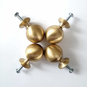 May include: Four gold-coloured spherical cabinet knobs with a brass finish arranged in a square. Each knob has a small, round base and a threaded screw for attachment. The text "AntiqKent" is visible on each knob.