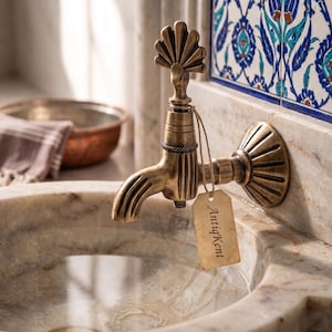 Op de afbeelding: Een wandkraan van messing met een verweerde afwerking, voorzien van een waaiervormige hendel en een decoratieve voet. Een klein, rechthoekig beige papieren label met de tekst AntiqKent is met een dun bruin touwtje aan de uitloop bevestigd.