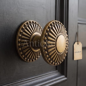 Può includere: Un pomello per porta rotondo in ottone con un motivo a raggiera e un bordo perlato. Il pomello presenta un disco centrale liscio ed è montato su una porta scura e strutturata. Un piccolo cartellino rettangolare beige con il testo AntiqKent pende.
