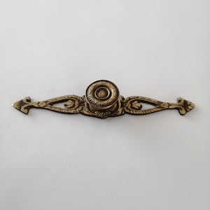 Puede incluir: Un tirador de cajón de metal dorado con un diseño ornamentado. El tirador tiene un pomo central con un patrón circular en relieve y una placa rectangular larga con detalles intrincados.