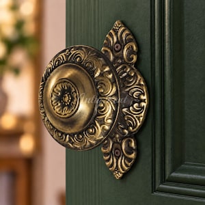May include: A round, antique brass door knob featuring intricate embossed scrollwork and a central floral motif. The knob is mounted on a dark green door with a decorative backplate that extends vertically, secured by two visible metal screws.