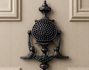 May include: A black metal door knocker featuring a central circular plate with a textured honeycomb pattern, flanked by two stylized bird figures at the base. The design includes a vertical structure with decorative finials and the text Antiq Kena.